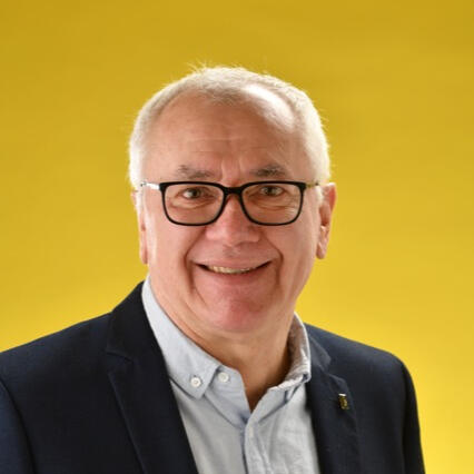 Photo de Bruno Mascle au format portrait sur fond jaune, chemise et veste de costume, visage souriant et lunettes.